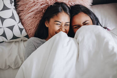 Two girls laughing while lying on bed during a sleepover. Top view of happy girls lying on bed together covering a blanket. - JLPSF20838