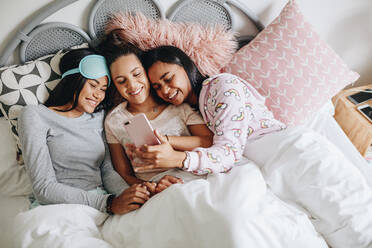 Draufsicht auf drei glückliche Mädchen, die auf ein Handy schauen, während sie auf dem Bett liegen und ein Selfie machen. - JLPSF20845