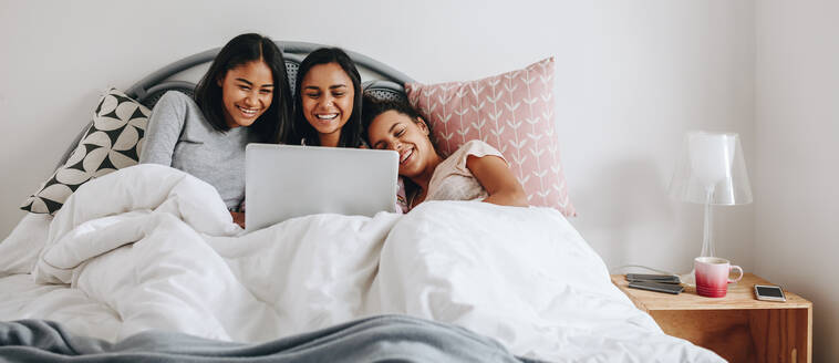 Three girls lying in bed covered in a blanket watching movie on a laptop. Girls having fun using a laptop lying on bed during a sleepover. - JLPSF20850