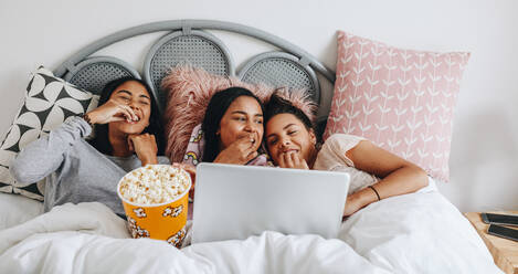 Mädchen, die eine Übernachtung haben, genießen einen Film auf dem Laptop mit Popcorns. Drei junge Mädchen, die Spaß haben, einen Film auf dem Laptop zu sehen und Popcorns in einer Wanne während einer Übernachtung zu essen. - JLPSF20852