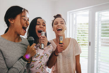 Three girls singing holding microphones standing in their room. Happy girls singing karaoke at a sleepover. - JLPSF20853