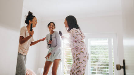 Drei Mädchen stehen auf einem Bett und halten Haarbürsten und eine Fernbedienung in der Hand und singen im Karaoke-Stil. Glückliche Mädchen bei einer Übernachtung, die das gemeinsame Singen genießen. - JLPSF20858