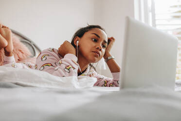 Junges Mädchen im Bett, das einen Film auf dem Laptop anschaut. Mädchen mit Laptop auf dem Bett liegend, während sie mit Kopfhörern Musik hört. - JLPSF20861