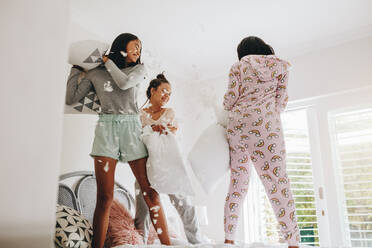 Fröhliche Mädchen spielen mit Kissen auf dem Bett während einer Übernachtung zu Hause. Drei junge Mädchen haben Spaß beim Kampf mit Kissen, die auf dem Bett stehen, wobei Federn herumfliegen. - JLPSF20867