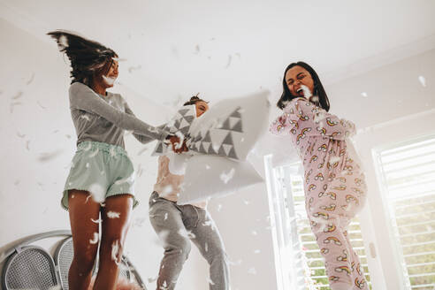 Three girls fighting with pillows standing on bed with feathers flying everywhere. Girls enjoying pillow fight standing on bed at a sleepover. - JLPSF20868