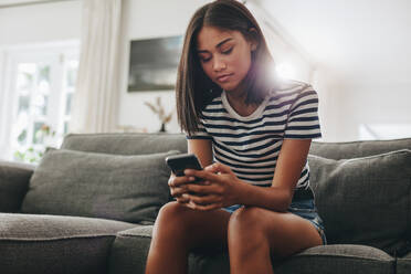 Teenage girl typing text message on a cell phone sitting at home. Young girl checking her mobile phone sitting on couch at home. - JLPSF20875