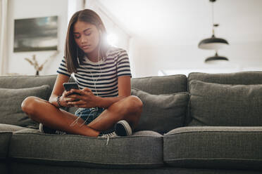 Young girl relaxing at home listening to music using earphone on a mobile phone. Teenage girl sitting on a couch at home enjoying music. - JLPSF20876