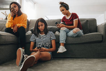 Three girls sitting at home listening to music independently using headphones. Friends sitting at home and checking their mobile phones. - JLPSF20877