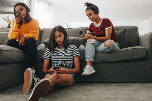 Drei Mädchen, die zu Hause sitzen und unabhängig voneinander mit Kopfhörern Musik hören. Freunde, die zu Hause sitzen und ihre Handys überprüfen. - JLPSF20877