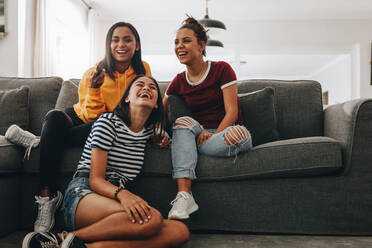 Three young girls sitting together at home watching television and laughing. Teenage girls at a sleepover having fun watching TV sitting in the living room. - JLPSF20878