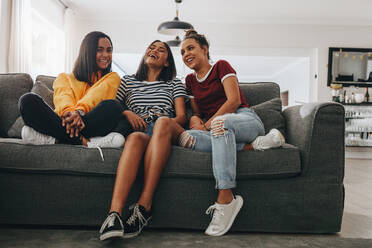 Three young girls sitting together on a couch in living room and laughing. Teenage girls having fun sitting together and talking at a sleepover. - JLPSF20879