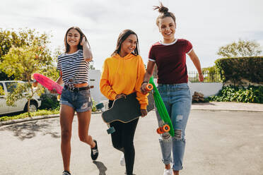 Aufgeregte Mädchen, die mit ihren Longboards und Skateboards auf der Straße laufen. Lächelnde Teenager-Mädchen, die sich an einem sonnigen Tag zum Skaten auf der Straße bereit machen. - JLPSF20882