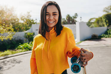 Fröhliches Teenager-Mädchen, das auf der Straße steht und ein Skateboard hält. Lächelndes Mädchen, das an einem sonnigen Tag draußen auf der Straße steht. - JLPSF20883