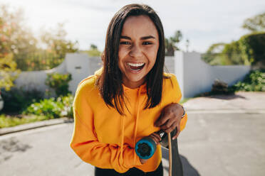 Close up of a laughing girl standing outdoors in the street. Girl standing in street on a sunny day holding a skateboard. - JLPSF20885