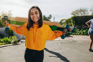 Lächelndes Teenager-Mädchen, das auf der Straße steht und ein Longboard auf den Schultern trägt. Junges Mädchen übt an einem sonnigen Tag auf der Straße Skaten. - JLPSF20887