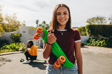 Ein lächelndes Teenager-Mädchen steht auf der Straße und hält ihr Skateboard in der Hand, während ihre Freundin im Hintergrund skatet. Junge Mädchen üben an einem sonnigen Tag das Skaten auf der Straße. - JLPSF20889