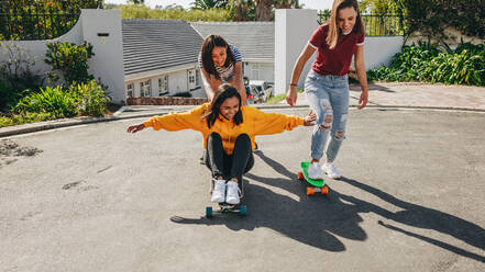 Three young girls having fun skating outdoors in street on a sunny day. Smiling girl pushing her friend who is sitting on a longboard from behind. - JLPSF20896