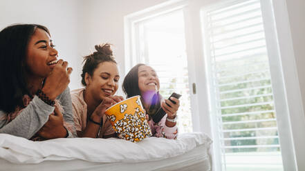 Drei junge Mädchen liegen auf dem Bett und essen Popcorn aus einer Wanne, während sie fernsehen. Die Mädchen genießen es, bei einer Übernachtung zu Hause fernzusehen. - JLPSF20897