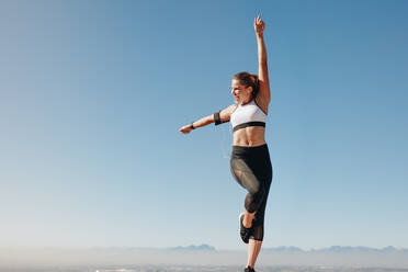 Fitness-Frau beim Workout auf einem Hügel stehend und Musik hörend. Fröhliche Frau in Fitnesskleidung beim Workout im Freien mit klarem blauen Himmel im Hintergrund. - JLPSF20901