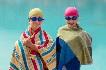 Two kids wrapped in towel standing by pool. Boy and girl in swimming goggles and cap after swimming training. - JLPSF20907