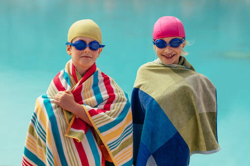 Zwei in ein Handtuch gewickelte Kinder stehen am Pool. Junge und Mädchen mit Schwimmbrille und Kappe nach dem Schwimmtraining. - JLPSF20907