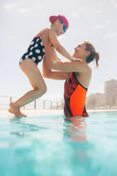 Woman taking a girl in the swimming pool from the edge of the pool. Mother and daughter at outdoor pool. - JLPSF20922
