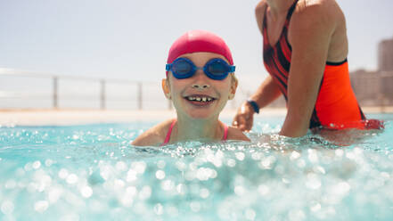 Niedliches Mädchen mit Badekappe und Schwimmbrille, das mit Hilfe seiner Mutter im Schwimmbad schwimmen lernt. Mädchen, das mit seiner Mutter Schwimmunterricht im Schwimmbad hat. - JLPSF20923