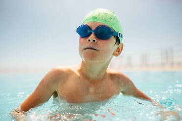 Junge mit Badekappe und Schwimmbrille schwimmt im Pool. Junge lernt schwimmen an einem Sommertag im Freibad. - JLPSF20932