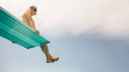 Niedriger Blickwinkel auf einen Jungen mit Schwimmbrille und Badekappe, der auf einem Sprungbrett gegen den hellen Himmel sitzt. Junge auf Sprungturm im Schwimmbad. - JLPSF20935