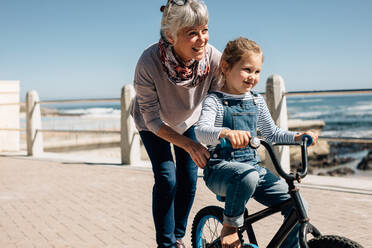 Mädchen, das mit Hilfe seiner Großmutter an der Strandpromenade versucht, Fahrrad zu fahren. Ältere Frau hält das Fahrrad, während ihre Enkelin das Fahren lernt. - JLPSF20936