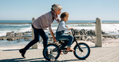 Seitenansicht eines Mädchens, das Fahrrad fährt, während seine Großmutter das Kind festhält. Eine ältere Frau bringt einem kleinen Mädchen auf der Straße am Strand das Fahrradfahren bei. - JLPSF20937