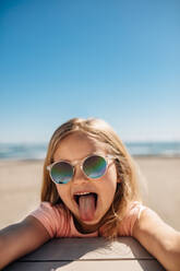 Verspieltes kleines Mädchen mit Sonnenbrille streckt ihre Zunge an der Strandpromenade heraus. Junges Mädchen, das an einem Sommertag am Strand lustige Gesichter macht. - JLPSF20941