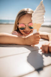 Beautiful girl holding a ice cream over a beach boardwalk. Young girl wearing sunglasses at the beach eating icecream on a summer day. - JLPSF20943