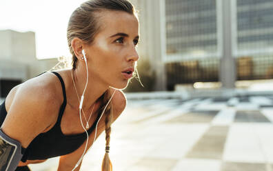 Weibliche Athletin entspannt sich beim Training im Freien stehend. Fitness-Frau macht Übungen im Freien und hört Musik mit Kopfhörern. - JLPSF20948