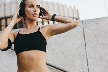 Close up of a fitness woman in training clothes listening to music on wireless headphones. Woman training outdoors listening to music looking away. - JLPSF20958