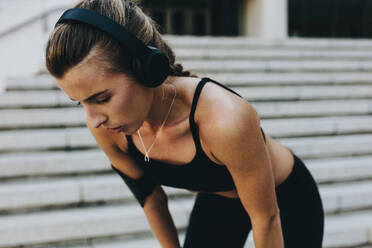 Fitness woman relaxing with hands on knees during workout standing outdoors. Fitness woman training outdoors listening to music wearing wireless headphones. - JLPSF20959