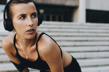 Close up of a female athlete relaxing during workout with hands on knees looking away. Fitness woman wearing wireless headphones training outdoors. - JLPSF20960