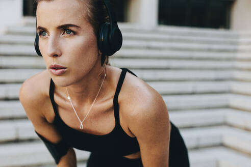 Close up of a woman athlete in training clothes standing outdoors with hands on knees. Fitness woman wearing wireless headphones relaxing while training. - JLPSF20961