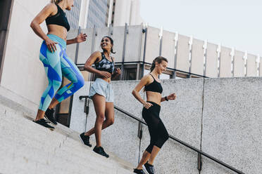 Weibliche Athleten, die beim Morgenlauf die Treppe eines Gebäudes hinunterjoggen. Eine Gruppe von drei Fitness-Frauen, die im Freien trainieren und die Treppe hinuntersteigen. - JLPSF20963