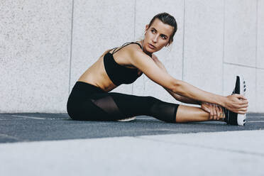 Fitness woman stretching her legs before workout sitting outdoors. Female runner doing stretching exercises listening to music. - JLPSF20965