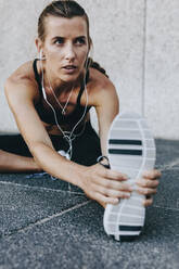 Close up of a woman athlete stretching her leg while listening to music using earphones. Fitness woman stretching forward and holding her shoe sitting outdoors. - JLPSF20966