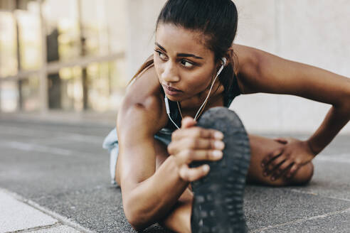 Fitness-Frau, die ihr Bein und ihren Rücken im Freien sitzend dehnt, Musik hört und Kopfhörer trägt. Fitness-Frau, die sich auf dem Boden sitzend nach vorne beugt und ihr Bein dehnt. - JLPSF20969