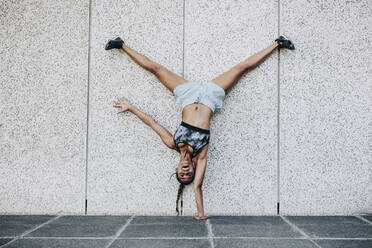 Female athlete in training clothes doing an acrobatic move turning upside down outdoors. Acrobatic fitness woman balancing upside down on one hand with legs in air. - JLPSF20972