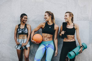 Lächelnde Sportlerinnen in Fitnesskleidung entspannen sich nach dem Training. Drei Fitness-Frauen stehen im Freien und halten Springseil, Basketball und Matte nach dem Training. - JLPSF20980