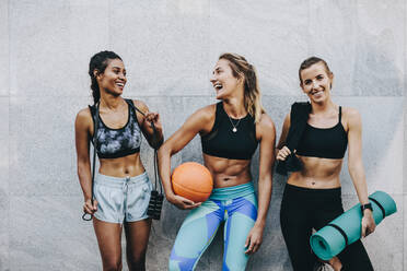 Glückliche Frauen in Fitness-Kleidung entspannen und Spaß haben stehend gegen eine Wand. Fitness-Frauen tragen Springseil Basketball und Trainingsmatte stehend nach dem Training. - JLPSF20981