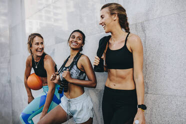 Drei Frauen in Fitnesskleidung, die nach dem Training im Freien an einer Wand stehen und miteinander reden. Glückliche Fitness-Frauen nach dem Training, die einen Basketball tragen und Seil springen. - JLPSF20985