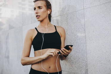 Women in fitness wear standing outdoors listening to music on mobile phone. Fitness woman standing against a wall listening to music using earphones. - JLPSF20988