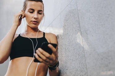 Fitness-Frauen entspannen sich nach dem Training und hören Musik auf dem Handy. Weibliche Athletin mit Kopfhörern steht während des Trainings an einer Wand. - JLPSF20989