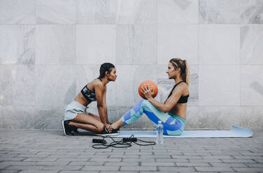 Frau in Fitnesskleidung macht Sit-Ups auf einer Übungsmatte, während ihre Freundin ihre Beine hält. Seitenansicht einer Fitnessfrau, die Sit-Ups macht und einen Basketball im Freien hält. - JLPSF20993