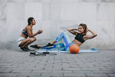 Fitness woman training outdoors sitting on exercise mat with skipping rope and basketball on the floor. Woman doing sit ups while her friend motivates and cheers her up. - JLPSF20994